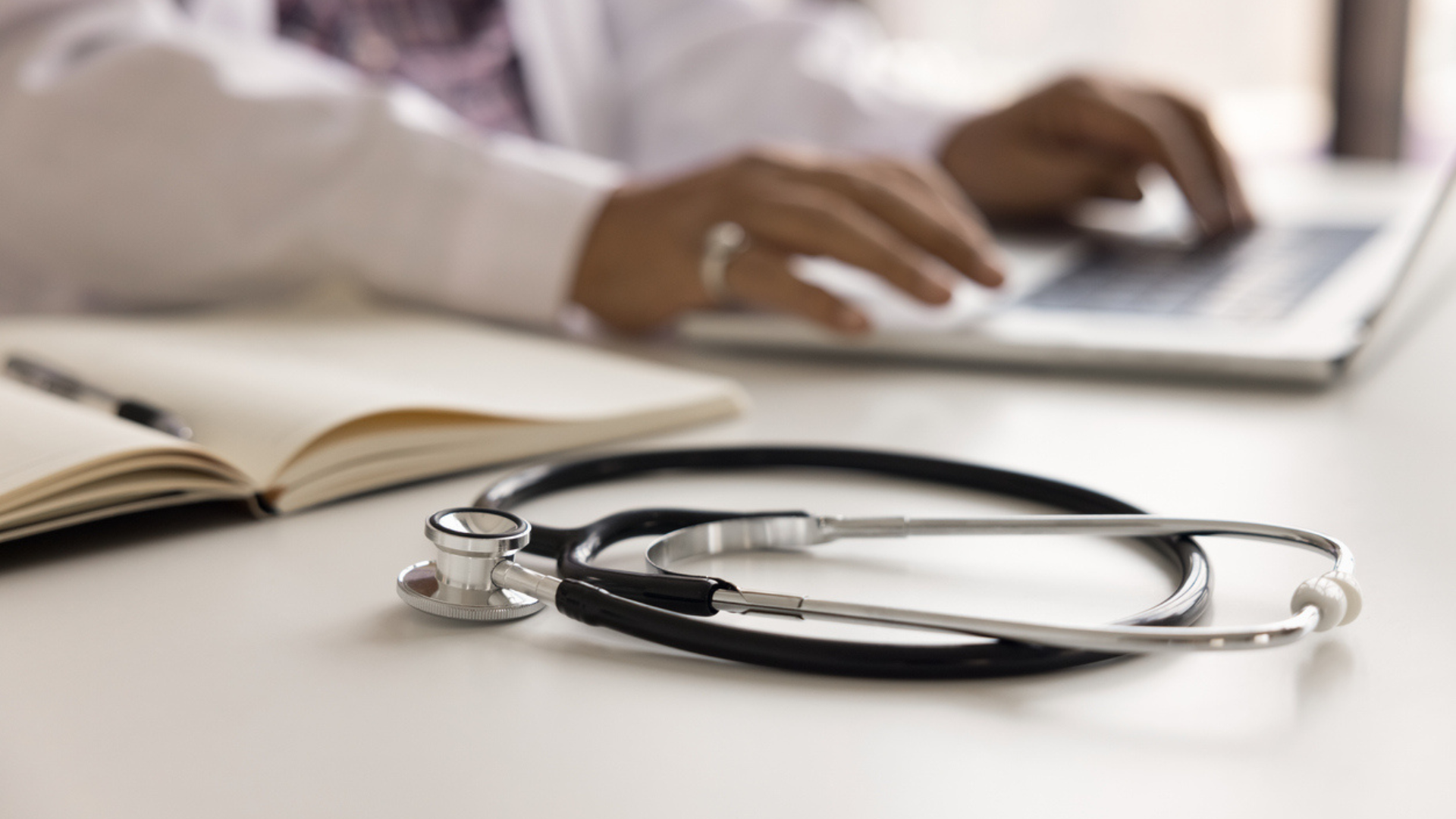Stethoscope on a desk with a person using a laptop in the background