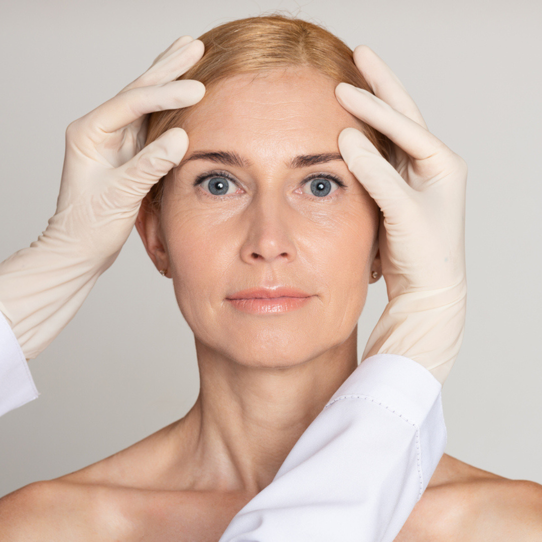 Woman with gloves on her hands examining her face against a neutral background
